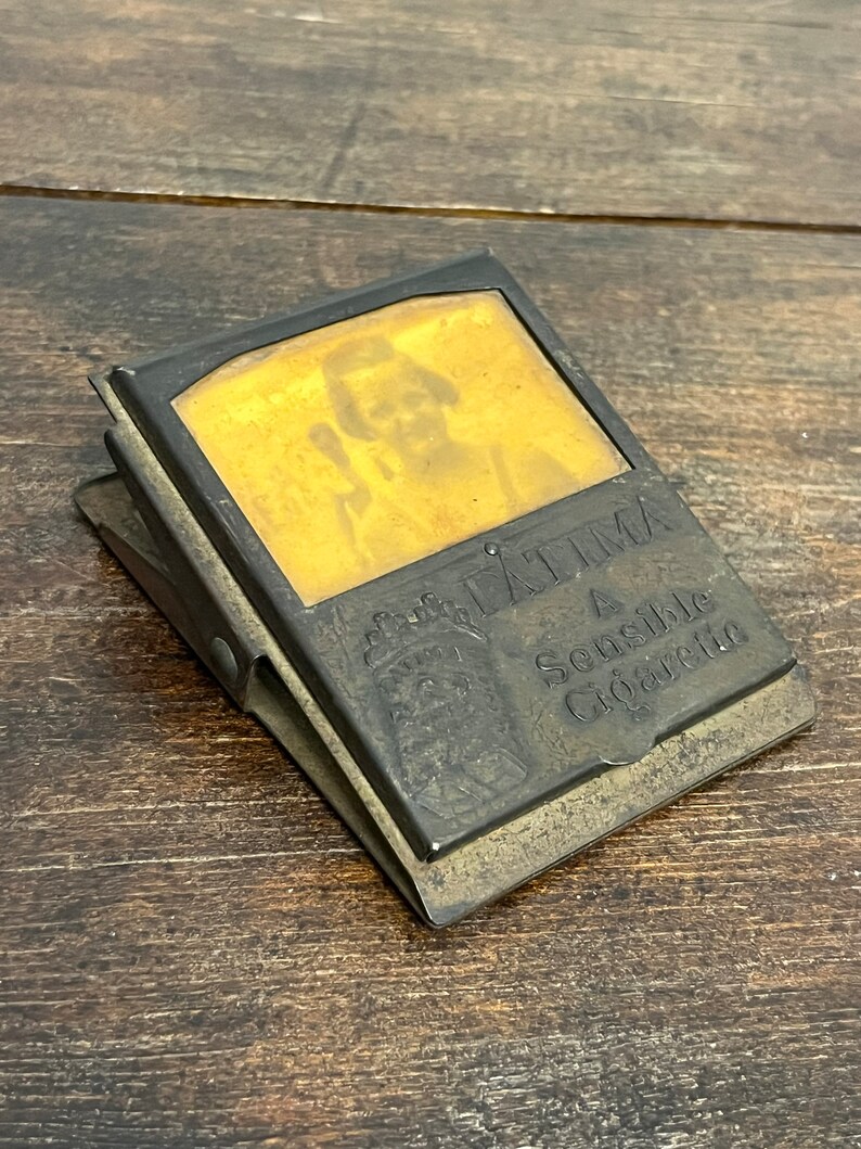 May include: Antique metal cigarette case with a hinged lid. The case has a rectangular shape with a faded image on the front. The text "FATIMA A Sensible Cigarette" is visible. The case is resting on a wooden surface.