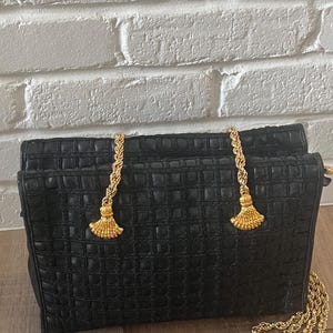 May include: A black handbag with a textured grid pattern across the surface. The bag features two gold-toned metal chain straps that attach to the top with decorative gold-toned fan-shaped tassels. A long gold-toned chain strap extends from the side.