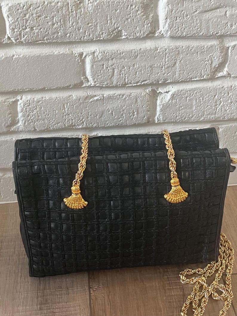 May include: A black handbag with a textured grid pattern across the surface. The bag features two gold-toned metal chain straps that attach to the top with decorative gold-toned fan-shaped tassels. A long gold-toned chain strap extends from the side.