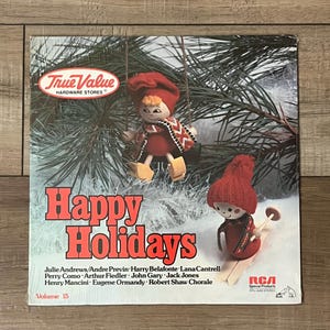 May include: A vinyl record cover titled Happy Holidays Volume 15, featuring two wooden dolls in red hats and scarves. One doll sits on a swing in pine branches, while the other skis on snow. The True Value Hardware Stores logo is in the top left corner.