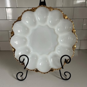 Puede incluir: Plato blanco para servir huevos con borde festoneado y ribete dorado, exhibido en un soporte de metal negro. El plato tiene 12 compartimentos individuales para huevos. Una pieza decorativa para servir.