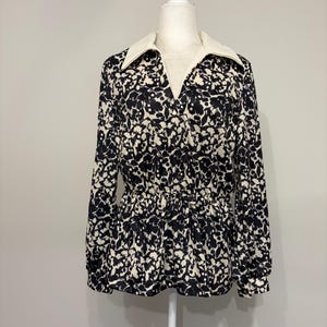 May include: A long-sleeved blouse featuring a black and white floral pattern, a solid white pointed collar, and a V-neckline. The garment includes a cinched waist with a drawstring detail, creating a gathered effect above the hemline.