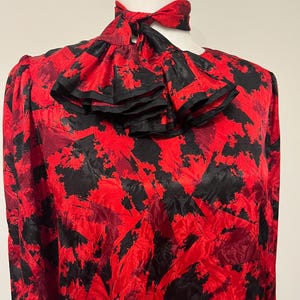 May include: A red and black patterned blouse with a high neck and a ruffled collar. The blouse has long sleeves and a matching necktie. The fabric has a textured appearance.