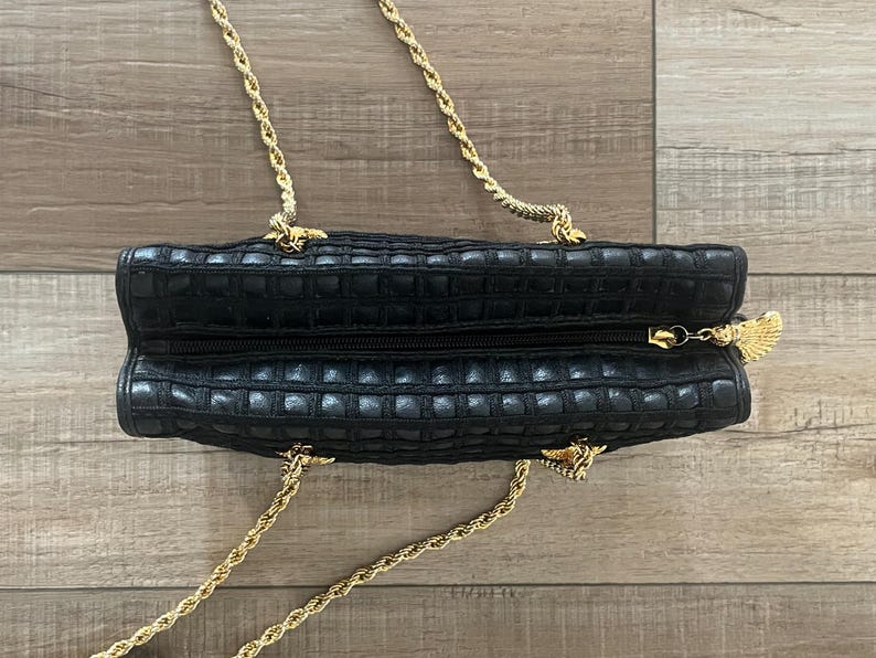 May include: A black rectangular handbag with a textured grid pattern and a top zipper closure. The bag features a gold-tone metal chain strap and a decorative gold-tone metal fan-shaped zipper pull attached to the zipper slider.