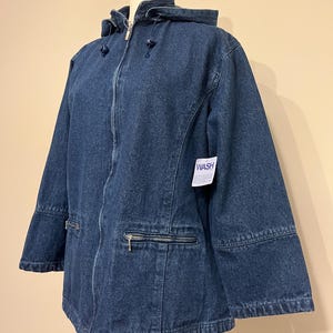 May include: A dark blue denim jacket with a hood, zipper closure, and two front pockets with zippers. The jacket has a tag that says "WASH". The jacket is displayed on a mannequin.