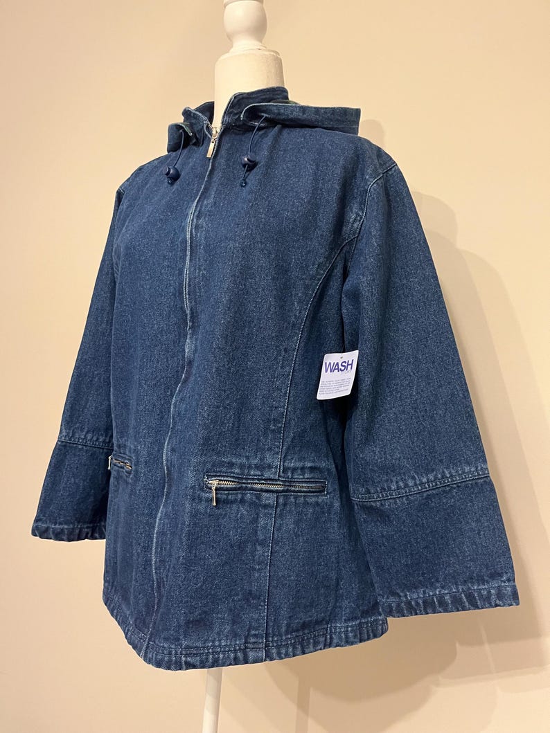 May include: A dark blue denim jacket with a hood, zipper closure, and two front pockets with zippers. The jacket has a tag that says "WASH". The jacket is displayed on a mannequin.