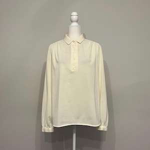 Puede incluir: Una blusa de color crema de manga larga con cuello y tapeta de tres botones. La blusa tiene un corte holgado con mangas con puños y se muestra en un maniquí sobre un fondo neutro. La tela parece ser un material ligero.