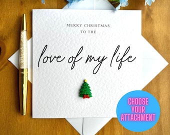 May include: White card with the text "Merry Christmas to the love of my life" and a small green Christmas tree with red ribbon. The card is on a wooden surface with blue flowers.