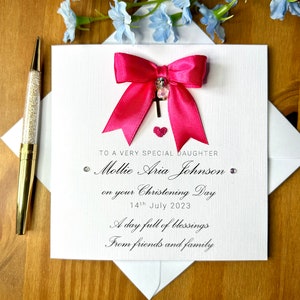 Puede incluir: Una tarjeta blanca con un lazo rosa y un dije de cruz plateada. La tarjeta dice "To a very special daughter Mollie Aria Johnson on your Christening Day 14th July 2023 A day full of blessings From friends and family"