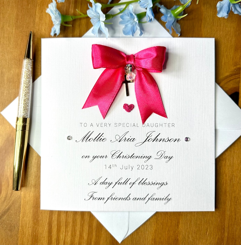 Puede incluir: Una tarjeta blanca con un lazo rosa y un dije de cruz plateada. La tarjeta dice "To a very special daughter Mollie Aria Johnson on your Christening Day 14th July 2023 A day full of blessings From friends and family"
