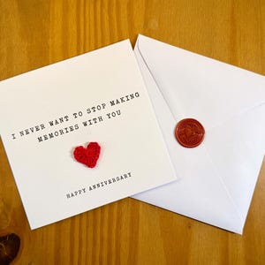 May include: A white card with the text "I NEVER WANT TO STOP MAKING MEMORIES WITH YOU" and a small red heart. Beside it is a white envelope with a red wax seal. The card reads "HAPPY ANNIVERSARY".