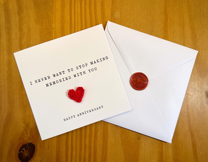 May include: A white card with the text "I NEVER WANT TO STOP MAKING MEMORIES WITH YOU" and a small red heart. Beside it is a white envelope with a red wax seal. The card reads "HAPPY ANNIVERSARY".