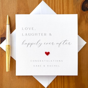 以下が含まれることがあります： 「LOVE, LAUGHTER & happily ever after」という言葉がエレガントな筆記体で書かれた白いウェディングカード。テキストの下には小さな赤いハートがあります。カードは白い封筒の上にあり、左側には金のペンがあります。カードには「おめでとうございます」と「GABE & RACHEL」の名前が書かれています。