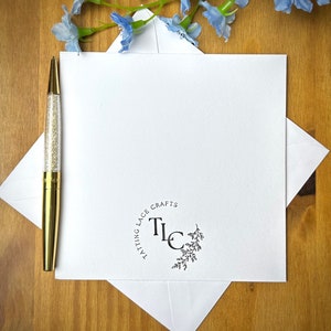 May include: A white stationery set with a gold pen and blue floral accents. The stationery includes a square card, an envelope, and a pen with a clear barrel filled with gold glitter. The card has a circular logo that reads "TATTTING LACE CRAFTS" with the letters "TLC".