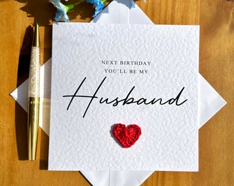 May include: White card with the text "Next birthday you'll be my Husband" in black script font. A small red crocheted heart is below the text.