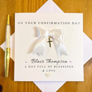 Può includere: Un biglietto di cresima bianco con un nastro bianco e un ciondolo a forma di croce dorata. Il biglietto recita "ON YOUR CONFIRMATION DAY" e include il nome "Blair Thompson" e il messaggio "A DAY FULL OF BLESSINGS & LOVE". C'è una penna d'oro a sinistra.