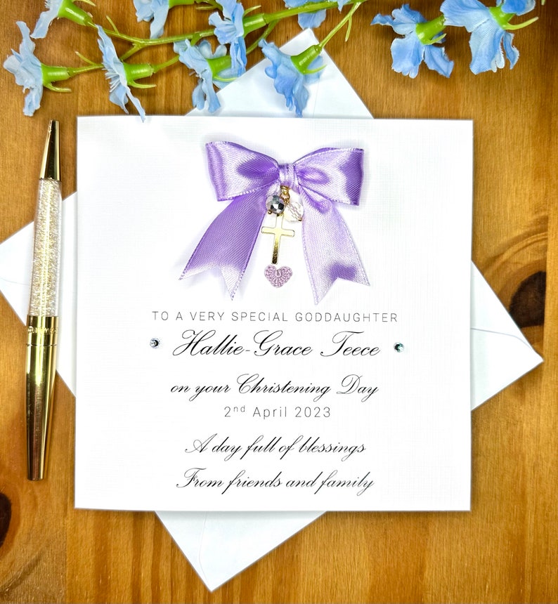 Puede incluir: Una tarjeta blanca con un lazo morado y un dije de cruz dorada. La tarjeta dice "To a very special goddaughter Hallie-Grace Teece on your Christening Day 2nd April 2023 A day full of blessings From friends and family".