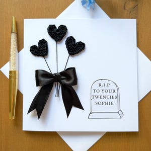 Op de afbeelding: Een witte wenskaart met een zwarte strik en drie zwarte gehaakte harten. De kaart heeft een grafsteenafbeelding met de tekst "R.I.P. TO YOUR TWENTIES SOPHIE". Links staat een gouden pen.