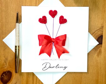 May include: A white card with a red bow and three red crocheted hearts. The card says "Happy Valentine's Day Darling".