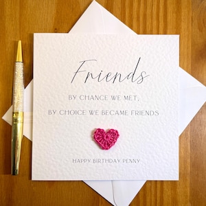 以下が含まれることがあります： 「Friends」の文字が書かれた白いグリーティングカード。テキストは「By chance we met, by choice we became friends」と書かれており、かぎ針編みのピンク色のハートが付いています。カードは白い封筒に入っており、金色のグリッターペンが付いています。