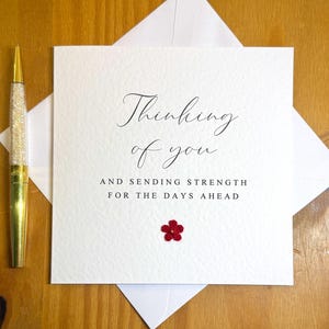 May include: A square white greeting card with the text Thinking of you and sending strength for the days ahead in black script and block lettering. A small red crocheted flower is attached below the text. A gold pen sits to the left.