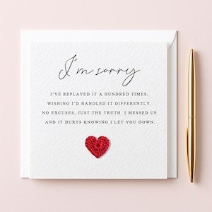 May include: A white card with the words "I'm sorry" in cursive. The card features a message of apology and a small, red crocheted heart. A gold pen is positioned beside the card on a pale pink surface.