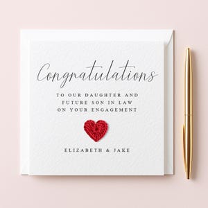 May include: A white engagement card with the word "Congratulations" in elegant script. The card includes the message "To our daughter and future son in law on your engagement" and a red crocheted heart. The names "Elizabeth & Jake" are printed at the bottom.