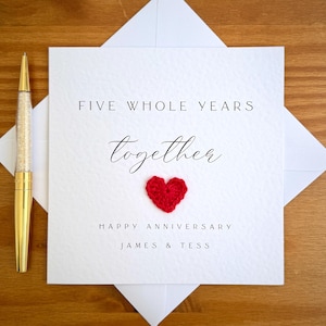 Puede incluir: Una tarjeta de aniversario blanca con el texto "FIVE WHOLE YEARS Together HAPPY ANNIVERSARY JAMES & TESS" y un pequeño corazón rojo. Un bolígrafo dorado con un cuerpo con incrustaciones de cristal descansa sobre una superficie de madera.