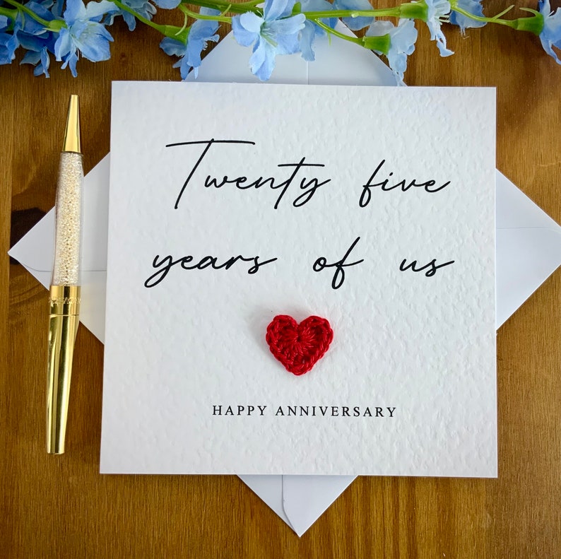 May include: A white card with black text that reads "Twenty five years of us" with a red crocheted heart in the center and the words "Happy Anniversary" below the heart.