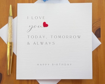 May include: A white greeting card with the text "I love you today, tomorrow & always" and a red heart. The card is on a white envelope, with a gold pen with a clear, glitter-filled barrel to the left. Blue flowers are at the top.