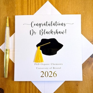May include: A white graduation card with the text "Congratulations Dr Blackshaw!" features a black mortarboard with a yellow tassel. The card also includes "PhD Organic Chemistry", "University of Bristol", and the year "2026". A gold pen with a clear, glitter-filled barrel is on the left.