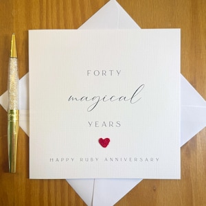 Könnte beinhalten: Eine quadratische, weiße Grußkarte mit strukturierter Oberfläche, die den schwarzen Text FORTY magical YEARS und HAPPY RUBY ANNIVERSARY sowie ein kleines rotes Herzsymbol zeigt. Neben der Karte liegt ein goldfarbener Stift mit kristallgefülltem Schaft.