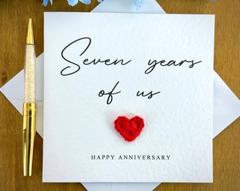 May include: A white card with the text "Seven years of us" in black cursive writing. A small red crocheted heart is below the text. The text "Happy Anniversary" is printed in black below the heart.