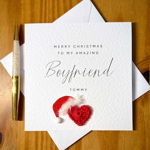 May include: A Christmas card with the message "MERRY CHRISTMAS TO MY AMAZING Boyfriend TOMMY". The card features a red Santa hat and a red crocheted heart. A gold pen with a glittery barrel is on the left.
