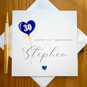 Könnte beinhalten: Eine weiße Geburtstagskarte mit einem blauen herzförmigen Ballon mit der Zahl 30. Die Karte trägt die Aufschrift "Happy 30th Birthday Stephen" in eleganter Schrift. Unten befindet sich ein kleines blaues Herz.