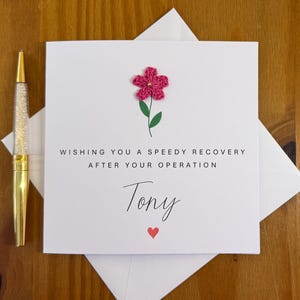 Peut inclure: Une carte de prompt rétablissement blanche avec le message "Wishing you a speedy recovery after your operation, Tony." Elle présente une fleur rose crochetée et un cœur rouge. Un stylo doré avec un corps à paillettes est à gauche.