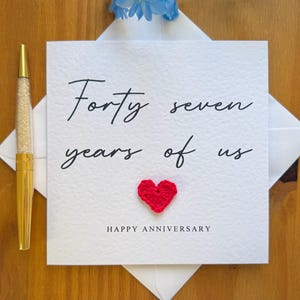 以下が含まれることがあります： 「Forty seven years of us」の黒い筆記体、小さな赤いハート、「Happy Anniversary」が印刷された白い記念日カード。クリスタルバレル付きのゴールドペンが左側にあります。