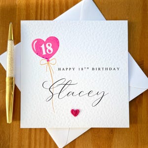 May include: A white birthday card with the text "Happy 18th Birthday Stacey." The card features a pink heart-shaped balloon with the number 18 inside. A small, red heart is at the bottom. A gold pen is on the left.