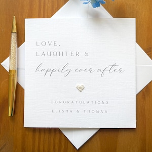 May include: A white wedding card with the words "Love, Laughter & Happily Ever After" printed in black script. The card features a small, textured heart and is accompanied by a gold pen with a glittery barrel. The card is on a wooden surface.