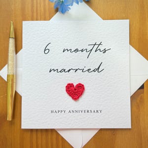 May include: A white anniversary card with the text "6 months married" and a red crocheted heart. The words "Happy Anniversary" are printed at the bottom. A gold pen with a glittery barrel is on the left.