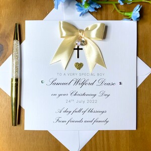 Puede incluir: Una tarjeta blanca con un lazo dorado y un dije de cruz. La tarjeta dice "To a very special boy Samuel Wilford Dease on your Christening Day 24th July 2022 A day full of blessings From friends and family"