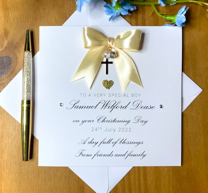 Puede incluir: Una tarjeta blanca con un lazo dorado y un dije de cruz. La tarjeta dice "To a very special boy Samuel Wilford Dease on your Christening Day 24th July 2022 A day full of blessings From friends and family"