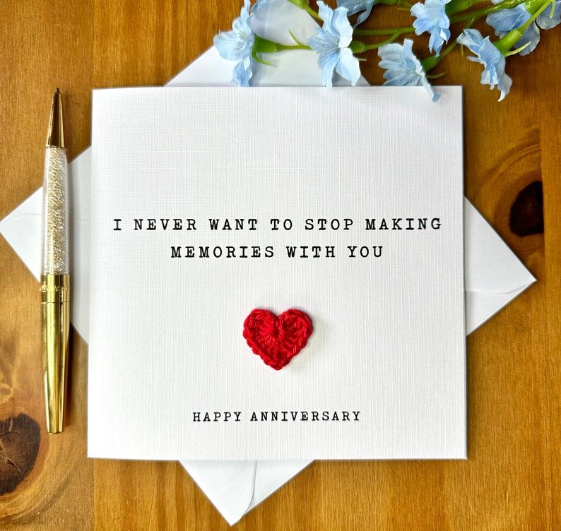 May include: A white anniversary card with the text "I NEVER WANT TO STOP MAKING MEMORIES WITH YOU" and "HAPPY ANNIVERSARY". A small, red, crocheted heart is centered below the text. A gold pen with a crystal barrel is on the left.