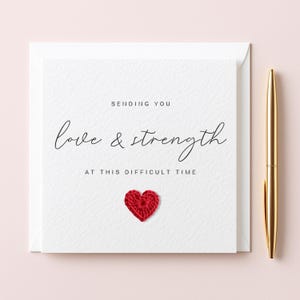 May include: A white card with the message "Sending You Love & Strength at this Difficult Time" in script. A small, red crocheted heart is below the text. A gold pen is positioned to the right of the card.