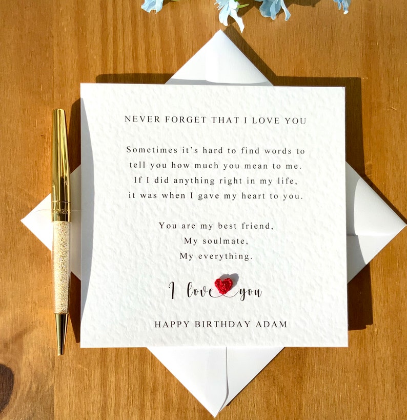 May include: A white birthday card with the message "NEVER FORGET THAT I LOVE YOU" and a loving poem. The card features a red heart and the words "I love you" and "HAPPY BIRTHAY ADAM". A gold pen is placed next to the card.