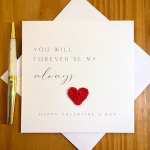 May include: A white Valentine's Day card with the message "YOU WILL FOREVER BE MY always" and a small red crocheted heart. A gold and crystal pen rests on the left side of the card. The card is inside a white envelope.
