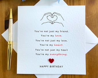 May include: A white card with a black line drawing of two hands forming a heart shape. The card has the text "You're not just my friend, You're my love. You're not just my love, You're my heart. You're not just my heart You're my everything." with the words "love", "heart", and "everything" in red. A small red heart is at the bottom of the card. The card also has the text "HAPPY BIRTHDAY" at the bottom.