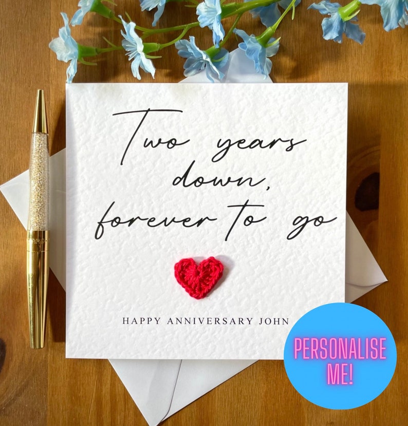 Pu&ograve; includere: Un biglietto bianco con il testo "Two years down, forever to go" scritto in corsivo nero. Un piccolo cuore rosso all'uncinetto si trova sotto il testo. Il biglietto dice anche "Happy Anniversary John" e ha un cerchio blu con il testo "Personalise Me!" in bianco.