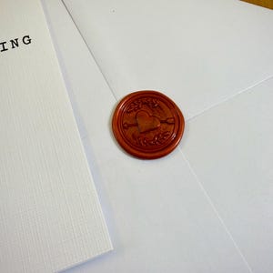 May include: A white envelope sealed with a round, reddish-brown wax seal. The seal features a heart pierced by an arrow, surrounded by decorative elements. A white card with the word "ING" is visible.