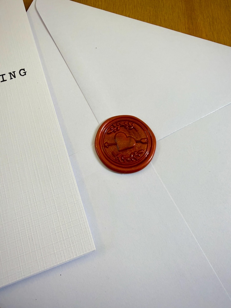 May include: A white envelope sealed with a round, reddish-brown wax seal. The seal features a heart pierced by an arrow, surrounded by decorative elements. A white card with the word "ING" is visible.